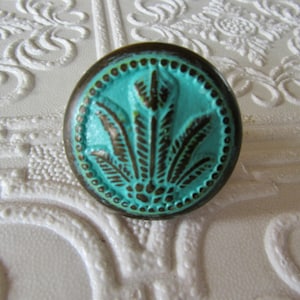 May include: A round, turquoise cabinet knob with a bronze-colored rim. The knob features a raised, stylized feather design in dark brown. The knob is set against a white, textured background with floral patterns.