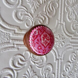 K&ouml;nnte beinhalten: Brauner Holzschrankknopf mit einem rosa-wei&szlig;en Blumendesign auf der Oberseite. Der Knopf hat einen wei&szlig;en Rand und ist vor einem wei&szlig;en, strukturierten Hintergrund platziert.
