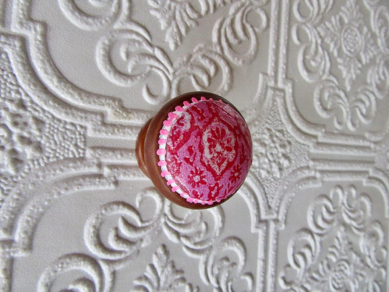 K&ouml;nnte beinhalten: Brauner Holzschrankknopf mit einem rosa-wei&szlig;en Blumendesign auf der Oberseite. Der Knopf hat einen wei&szlig;en Rand und ist vor einem wei&szlig;en, strukturierten Hintergrund platziert.