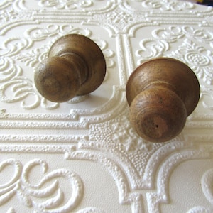 May include: Two antique wooden knobs with a rounded top and a smaller, spherical base. The knobs are a warm brown color and appear to be made of solid wood. They are set against a white, textured background with a repeating floral pattern.