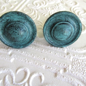May include: Two circular cabinet knobs with a distressed teal and dark green finish. Each knob features a raised center dome surrounded by a flat, circular rim, creating a layered, stepped profile with a textured, weathered appearance.