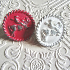 Puede incluir: Dos pomos de armario redondos decorativos. Uno es rojo con un jinete plateado sobre un caballo, y el otro es blanco con un jinete plateado sobre un caballo. Ambos pomos tienen un borde en forma de cuerda y están sobre una superficie blanca texturizada.