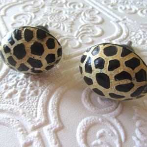 Pode incluir: Dois puxadores de armário ovais com um padrão de girafa preto e dourado. Cada puxador tem um pequeno caule de metal. O fundo é uma superfície branca texturizada.