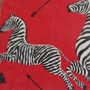 May include: A square coaster featuring a vibrant red background with two black and white striped zebras in motion. One zebra is positioned horizontally in the center, while the other is vertical on the right. Two thin black arrows point across the surface.