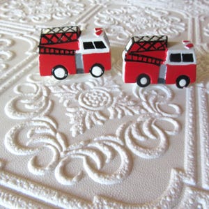 Pu&ograve; includere: Due figurine di camion dei pompieri rosse e bianche in miniatura con dettagli neri, tra cui scale e ruote. Le figurine sono poste su uno sfondo bianco strutturato, creando una composizione giocosa.