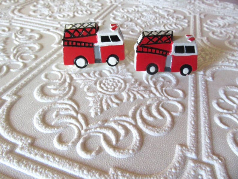 Pu&ograve; includere: Due figurine di camion dei pompieri rosse e bianche in miniatura con dettagli neri, tra cui scale e ruote. Le figurine sono poste su uno sfondo bianco strutturato, creando una composizione giocosa.