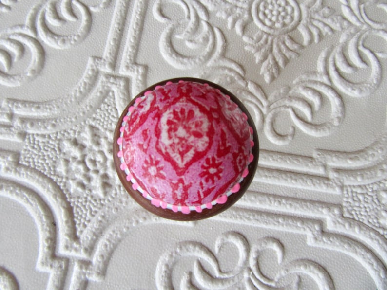 May include: A round, decorative cabinet knob with a pink and white floral design on a dark brown base. The knob has a white border and is set against a textured white background.
