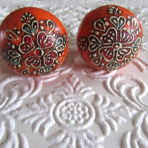 May include: Two round orange cabinet knobs featuring a hand-painted floral pattern in black, white, and red. The spherical knobs have a smooth finish and display a symmetrical, intricate design of petals and leaves across their surface.