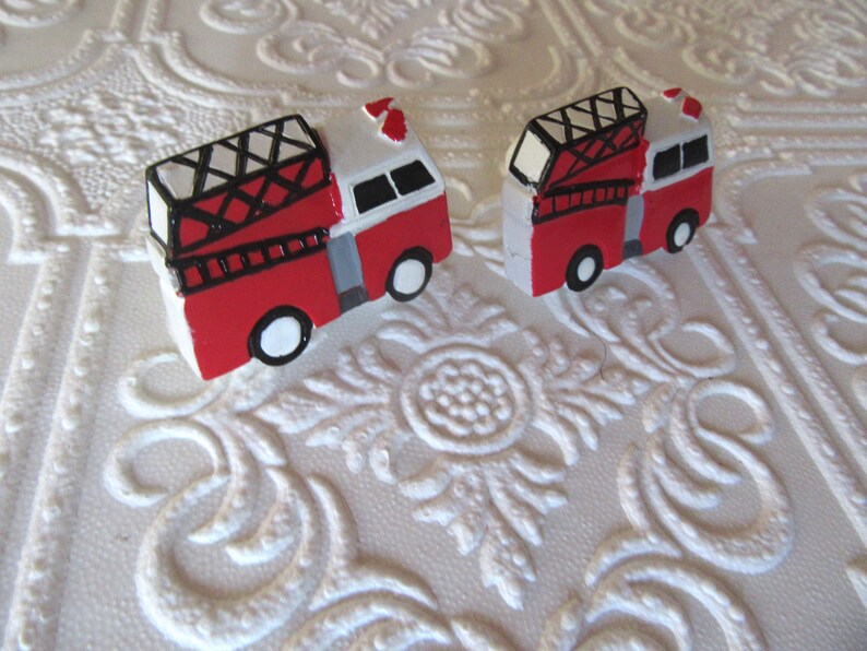 Pu&ograve; includere: Due camion dei pompieri in miniatura dipinti a mano. Ogni camion &egrave; rosso con tetto bianco, scala nera e ruote bianche e nere. Lo sfondo &egrave; una superficie bianca strutturata con un motivo floreale.