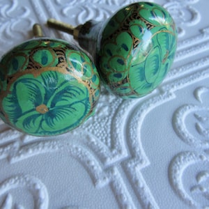 May include: Two decorative cabinet knobs with a green and gold floral design. The knobs are painted with a light green base color and feature a darker green floral pattern with gold accents. The knobs are on a white textured surface.