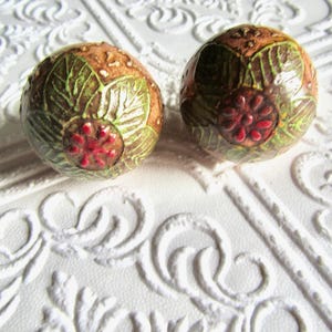 May include: Two spherical decorative items. They feature a brown base, green leaf patterns, and a red floral centre. The items rest on a white, textured surface with a repeating floral design. The items appear to be handmade.