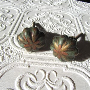 May include: Two decorative cabinet knobs with a floral design. The knobs are a mix of green, gold, and copper colors. The knobs are on a white textured surface.