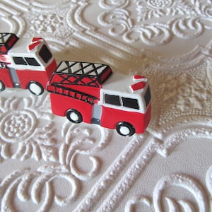 Pu&ograve; includere: Due camion dei pompieri rossi e bianchi in miniatura con dettagli neri, tra cui scale e finestre. I giocattoli sono su una superficie bianca strutturata con motivi floreali.