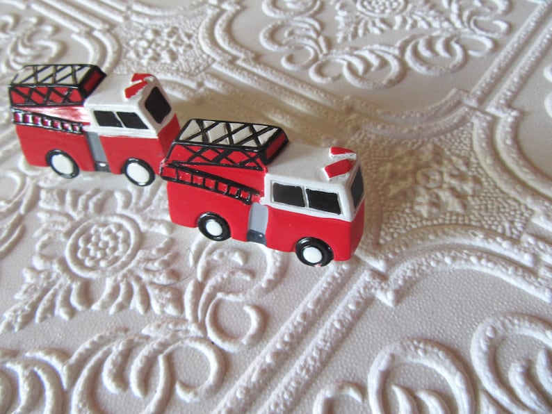 Pu&ograve; includere: Due camion dei pompieri rossi e bianchi in miniatura con dettagli neri, tra cui scale e finestre. I giocattoli sono su una superficie bianca strutturata con motivi floreali.