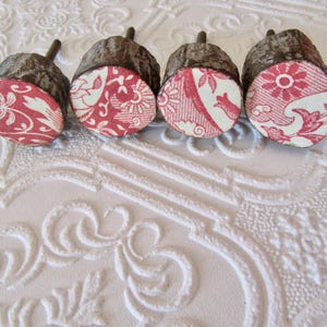 May include: Four decorative acorn-shaped objects. The tops are textured brown, and the bottoms feature red and white patterned fabric. The patterns vary, including floral and geometric designs. The acorns are arranged in a row.
