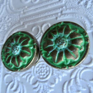 May include: Two antique-style, round cabinet knobs. Each knob features a raised floral design with a green and bronze finish. The knobs are set against a white, textured background.