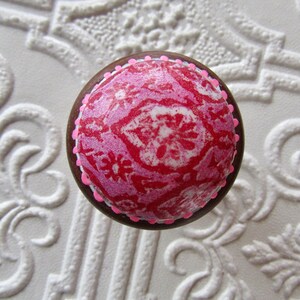 K&ouml;nnte beinhalten: Runder Schrankknopf mit braunem Rand und einem rosa-roten Muster auf der Oberseite. Der Knopf hat einen rosa gepunkteten Rand und ist vor einem wei&szlig;en, strukturierten Hintergrund platziert.
