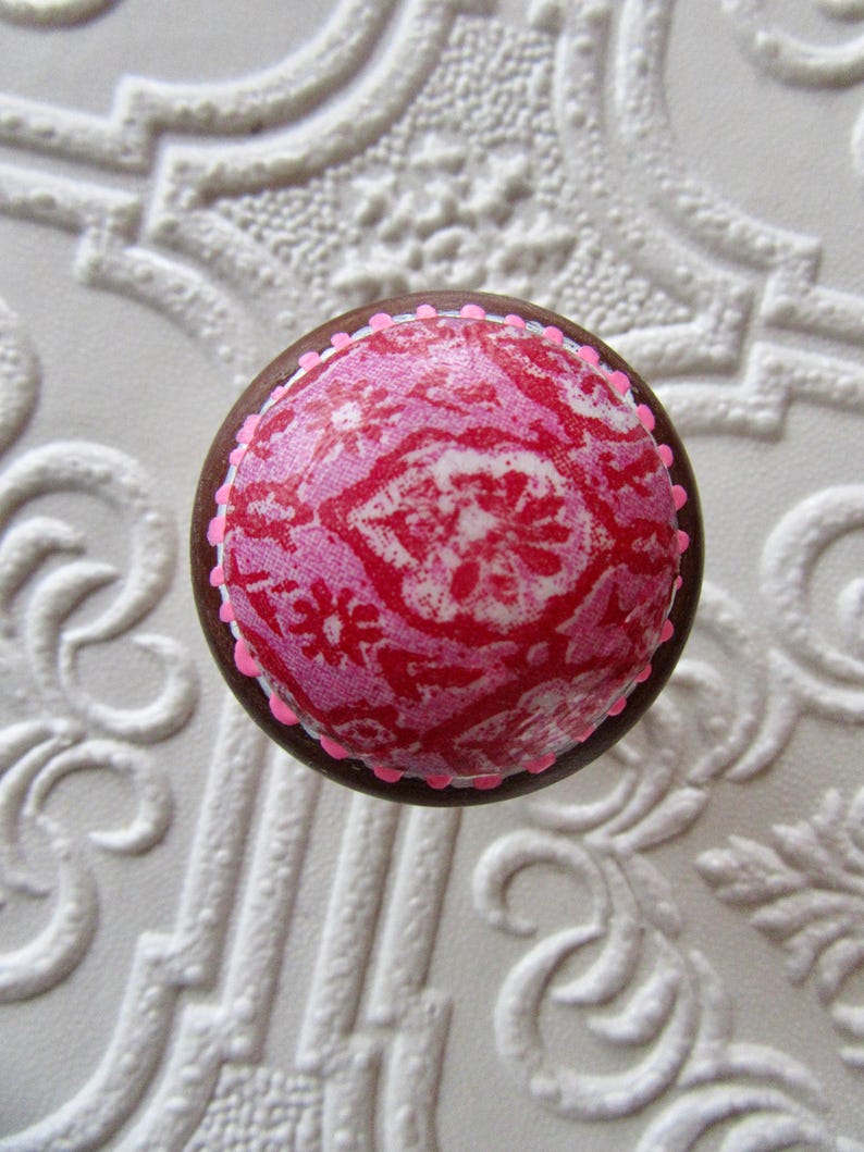 K&ouml;nnte beinhalten: Runder Schrankknopf mit braunem Rand und einem rosa-roten Muster auf der Oberseite. Der Knopf hat einen rosa gepunkteten Rand und ist vor einem wei&szlig;en, strukturierten Hintergrund platziert.