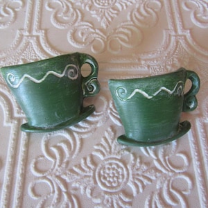 May include: Two small, green ceramic teacups with a white wavy design around the rim. Each cup has a handle and sits on a small saucer. The cups are displayed on a textured, off-white surface.