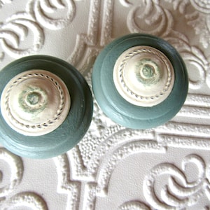 May include: Two round, teal-colored cabinet knobs with off-white, decorative centers. The centers have a circular, rope-like design around a central, floral-like detail. The knobs are set against a textured, white background.