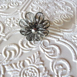 May include: A silver filigree flower brooch with a central bead. The brooch features delicate, openwork petals and is set against a textured, white background with embossed floral patterns. A vintage accessory.