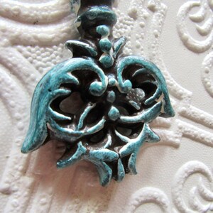 Op de afbeelding: Close-up van een decoratieve, sierlijke kastknop met een turquoise en bronzen afwerking. De knop heeft een gedetailleerd, symmetrisch ontwerp met krullende elementen en een schroefdraad. De achtergrond is een witte textuur.