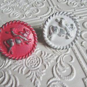 May include: Two decorative round buttons. One is a vibrant red with a silver horse and rider design, framed by a silver rope-like border. The other button is white with a similar design and a gray border.