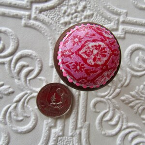 K&ouml;nnte beinhalten: Ein runder, dekorativer M&ouml;belknopf mit einem rosa-roten Blumenmuster. Der Knopf hat einen braunen Rand und eine rosa-wei&szlig;e Verzierung. Eine M&uuml;nze liegt neben dem Knopf zum Gr&ouml;&szlig;envergleich. Der Knopf liegt auf einer wei&szlig;en, strukturierten Oberfl&auml;che.