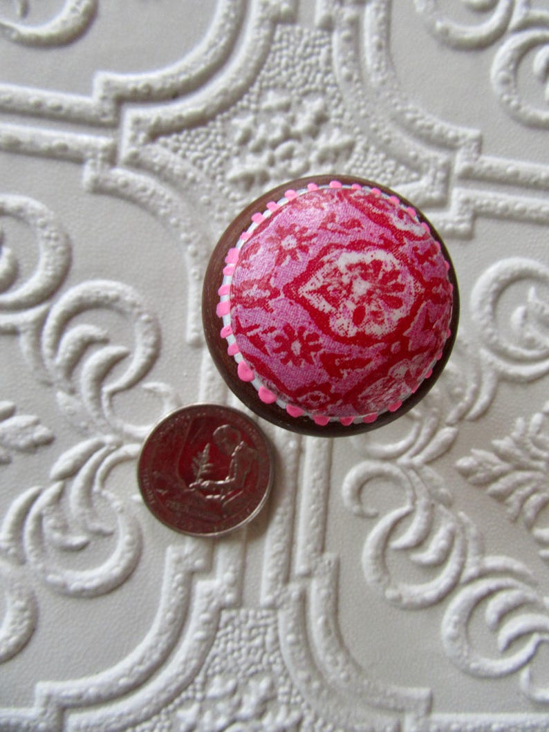 K&ouml;nnte beinhalten: Ein runder, dekorativer M&ouml;belknopf mit einem rosa-roten Blumenmuster. Der Knopf hat einen braunen Rand und eine rosa-wei&szlig;e Verzierung. Eine M&uuml;nze liegt neben dem Knopf zum Gr&ouml;&szlig;envergleich. Der Knopf liegt auf einer wei&szlig;en, strukturierten Oberfl&auml;che.