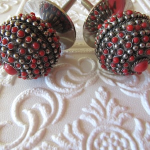 May include: Two decorative cabinet knobs with a silver-colored metal base and a spherical design. The spheres are covered in small red and silver-colored beads and larger red accents. The knobs are on a white, textured surface.