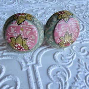 May include: Two decorative, round objects with a floral design. The objects have a pink center with a yellow flower and green and gold accents. They are set on a white, textured surface.