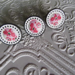 Op de afbeelding: Drie decoratieve kastknoppen. Elke knop heeft een wit midden met een roze bloemenontwerp, omgeven door een zwarte rand en een ring van kleine gaatjes. De knoppen zijn geplaatst tegen een getextureerde, grijze achtergrond.