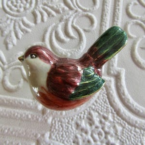 May include: A small ceramic bird figurine featuring a glossy finish with a cream-coloured chest, a deep reddish-brown head and body, and dark green accents on the wings and tail feathers. The bird is depicted in a side profile view.
