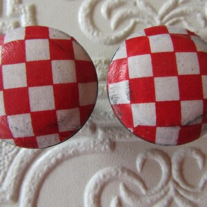 May include: Two circular buttons featuring a red and white checkered pattern. The surface of each button has a smooth, slightly glossy finish with visible texture and minor signs of wear along the edges of the red and white squares.