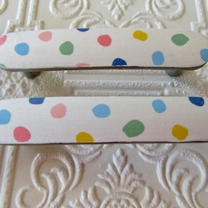 Può includere: Due maniglie per cassetti allungate con base bianca e un motivo a pois colorati. I pois sono nelle tonalità azzurro, blu scuro, rosa, giallo e verde. Ogni maniglia presenta una forma rettangolare arrotondata e liscia con fissaggi in metallo.