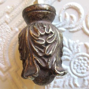 May include: A decorative furniture knob with a dark brown finish and gold accents. The knob features an ornate, leaf-like design. The background is a white, textured surface with a floral pattern.