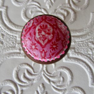 K&ouml;nnte beinhalten: Runder Schrankknopf mit einem rosa-wei&szlig;en Blumenmuster. Der Knopf hat eine braune Holzbasis und ein rosa-wei&szlig;es Design. Er befindet sich vor einem wei&szlig;en, strukturierten Hintergrund mit einem dekorativen Muster.