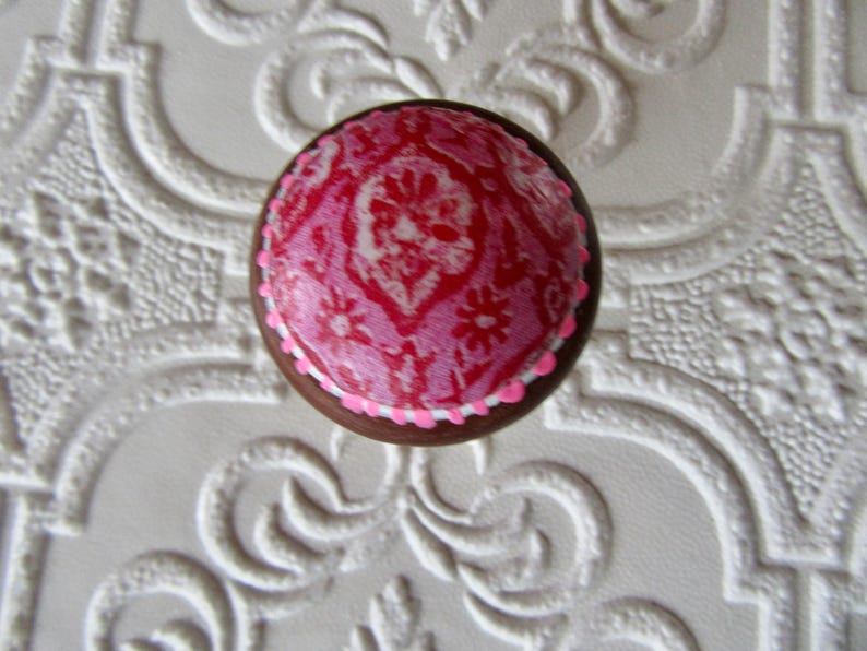 K&ouml;nnte beinhalten: Runder Schrankknopf mit einem rosa-wei&szlig;en Blumenmuster. Der Knopf hat eine braune Holzbasis und ein rosa-wei&szlig;es Design. Er befindet sich vor einem wei&szlig;en, strukturierten Hintergrund mit einem dekorativen Muster.