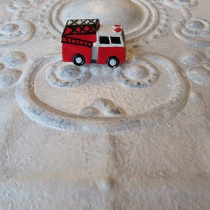 Pu&ograve; includere: Un piccolo camion dei pompieri giocattolo rosso e bianco con ruote nere e una scala nera. Il giocattolo &egrave; posto su uno sfondo strutturato bianco sporco con motivi ornamentali in rilievo.