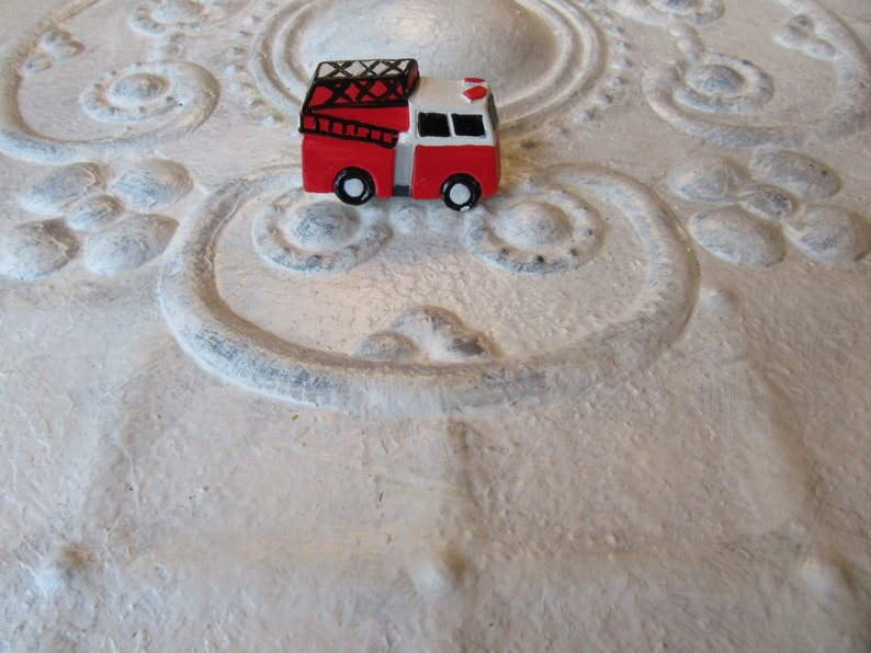 Pu&ograve; includere: Un piccolo camion dei pompieri giocattolo rosso e bianco con ruote nere e una scala nera. Il giocattolo &egrave; posto su uno sfondo strutturato bianco sporco con motivi ornamentali in rilievo.