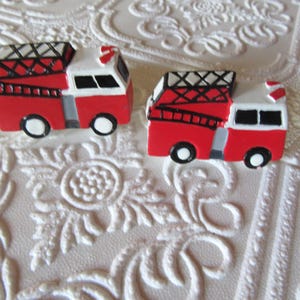 May include: Two miniature red and white fire trucks with black detailing and wheels. The trucks have ladders and a roof rack. The background is a white, textured surface with a floral pattern.