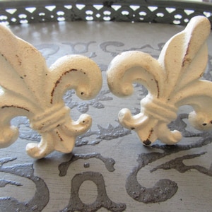 May include: Two off-white fleur-de-lis cabinet knobs with a distressed finish. The decorative hardware features a classic design with a slightly aged appearance, perfect for adding a touch of vintage charm to furniture or decor.