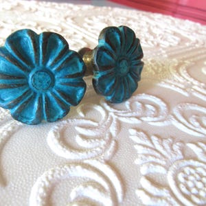 Peut inclure: Deux boutons de meuble turquoise en forme de fleur avec des accents sombres. Les boutons sont en métal et ont une finition vieillie. Ils sont posés sur un fond blanc texturé avec un motif floral. Quincaillerie décorative.