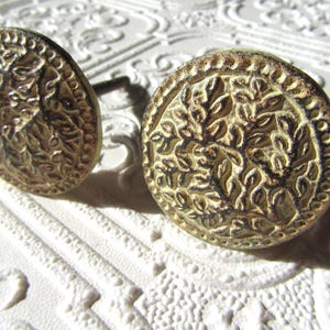 Może przedstawiać: Dwa okrągłe gałki do szafek w stylu antycznym z postarzanym złotym wykończeniem. Każda gałka ma szczegółowy, wypukły wzór kwiatowy wewnątrz obramowania z koralików. Gałki są umieszczone na teksturowanej białej powierzchni, tworząc estetykę vintage.