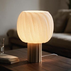 Può includere: Una lampada da tavolo con un paralume rotondo in vetro bianco smerigliato con una sottile trama a coste. La lampada ha una base cilindrica con un motivo a coste verticali abbinato ed emette una luce calda e soffusa.