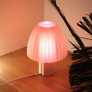 Könnte beinhalten: Eine rosa pilzförmige Lampe mit geripptem Lampenschirm und quadratischem Sockel. Die Lampe ist beleuchtet und strahlt sanft. Die Lampe steht auf einer Holzoberfläche.