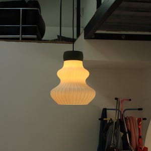 Op de afbeelding: Een moderne hanglamp met een crèmekleurige, geribbelde glazen kap en een donkergroene bovenkant. Het licht hangt aan het plafond met een zwart snoer. De armatuur is verlicht en geeft een warme gloed.
