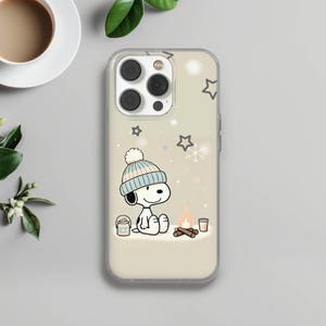May include: A clear phone case with a cartoon Snoopy design, wearing a winter hat and sitting by a campfire. The design includes stars and snowflakes. A cup of coffee and greenery are in the background.
