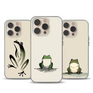 May include: Three phone cases with frog illustrations. The cases have a cream-colored background. One case features a black and green frog in a stylized pose. The other two cases have green frogs with white bellies in different poses.