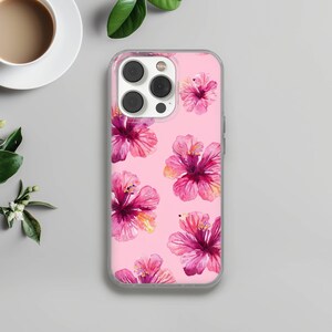 May include: A clear phone case with a pink background featuring a pattern of pink and magenta hibiscus flowers. The flowers have yellow stamens and are arranged in a repeating design. The case has a camera cutout.